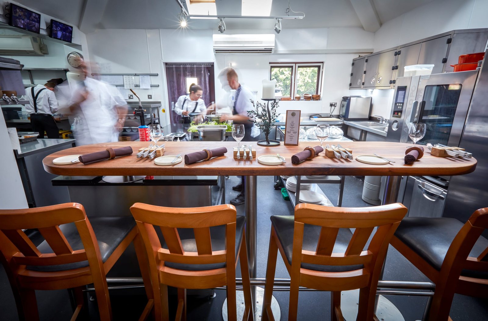 Four chefs move through a bright stainless-steel kitchen behind an empty wooden counter set with plates, glasses and folded napkins.