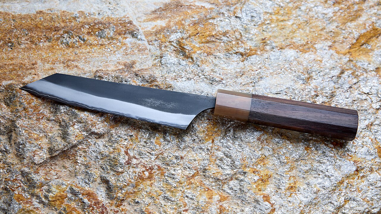 A dark-finished Japanese kiritsuke-style petty knife with a pointed tip and wooden handle resting on textured stone.