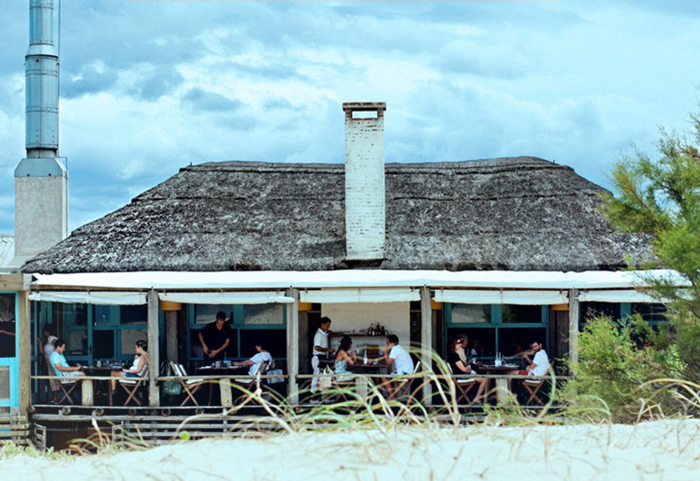 The open-sided dining terrace of Parador La Huella, with wooden tables beneath a thatched roof and beach grasses in the foreground.