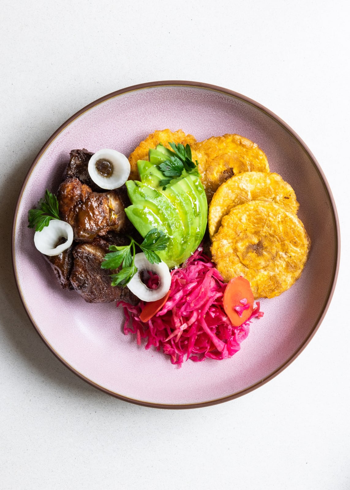 Overhead view of dark fried pork griyo with golden plantain rounds, sliced avocado, vivid pink pikliz and white onion on a pink plate.