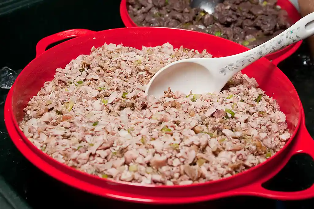 A wide red serving bowl filled with chopped chicken kelaguen and a white serving spoon, with a bowl of beef kelaguen behind it.