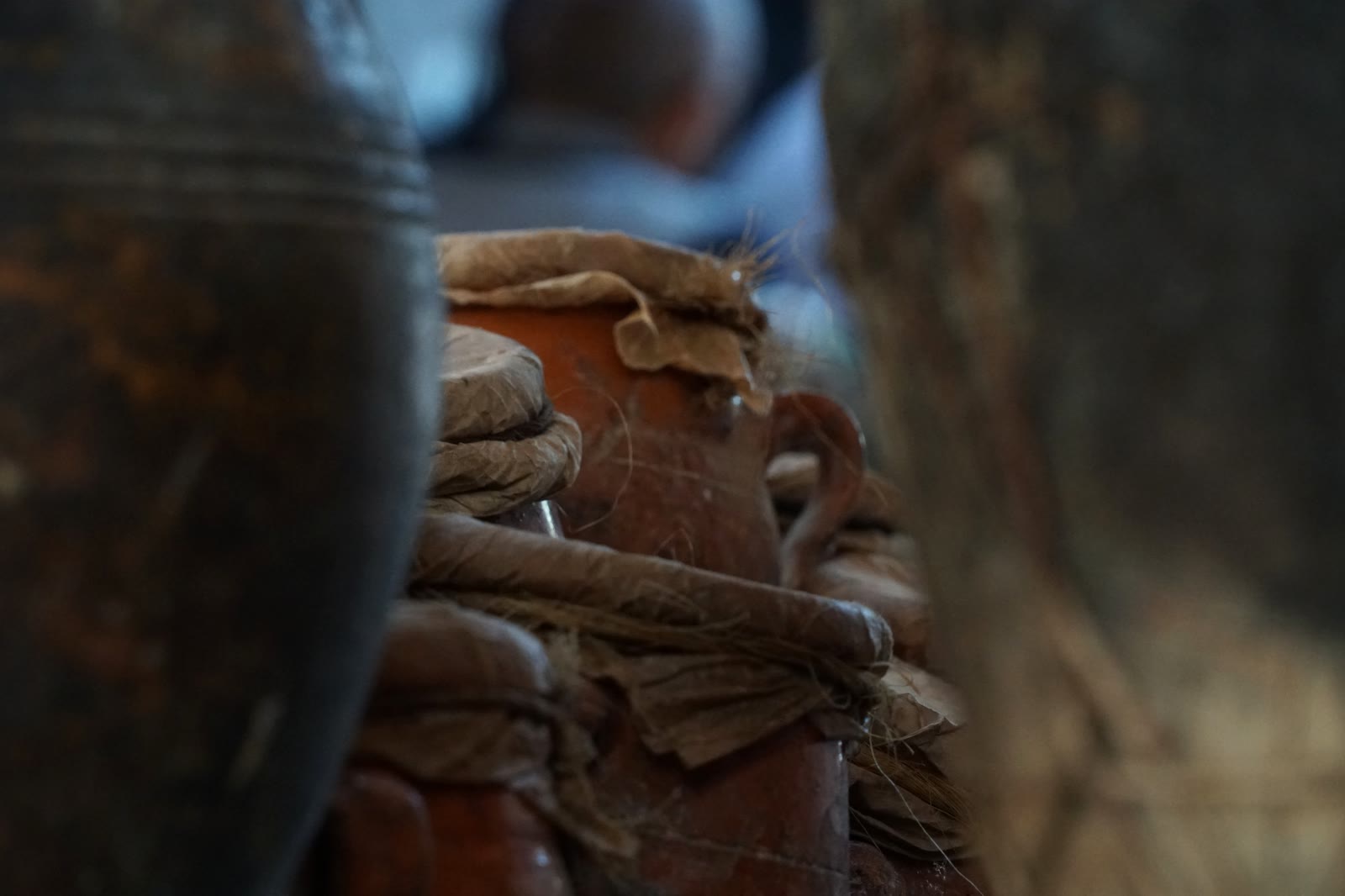 Paper-sealed terracotta tanjia jars stacked in a Marrakech souk, with their tied fiber and narrow necks visible between blurred shelves.