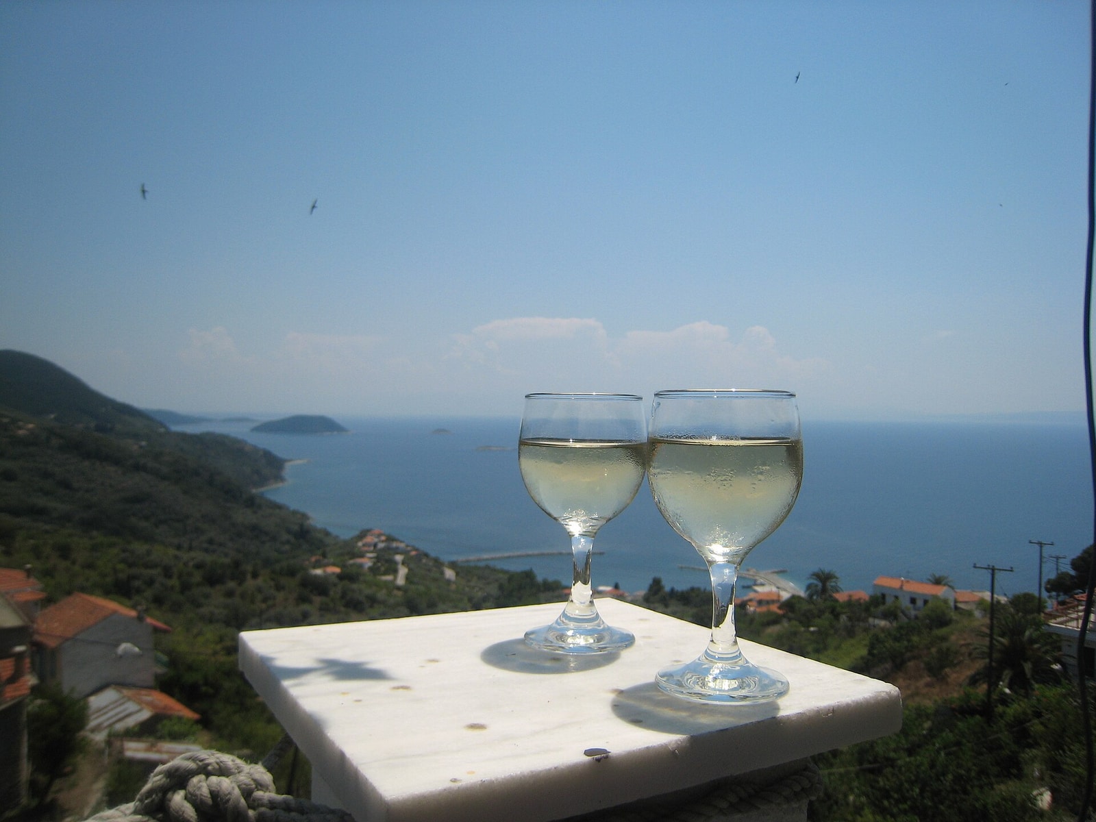 Two glasses of pale retsina on a white table overlooking a sunlit coastal landscape.