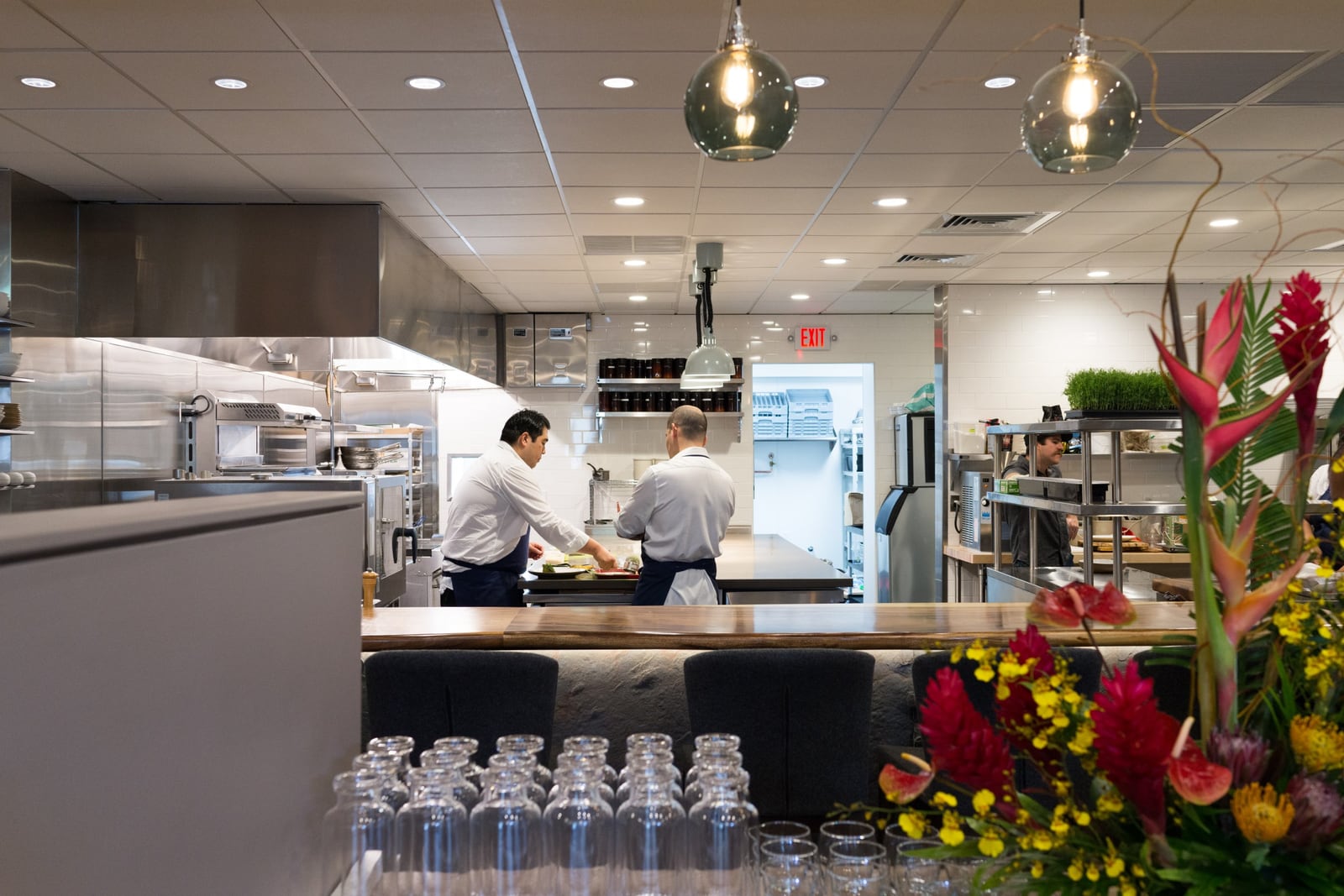 Two chefs in white jackets plate food in a bright open kitchen behind a wooden counter, with tropical flowers in the foreground.