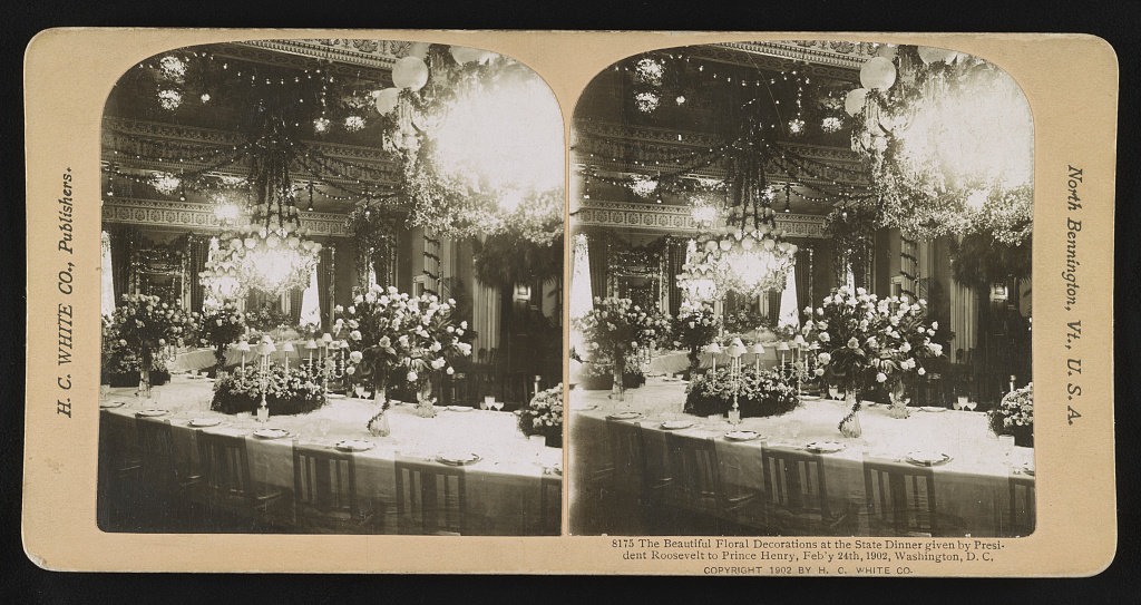 A sepia stereograph shows a crescent-shaped White House state-dinner table covered with flowers, glasses and place settings beneath chandeliers in 1902.