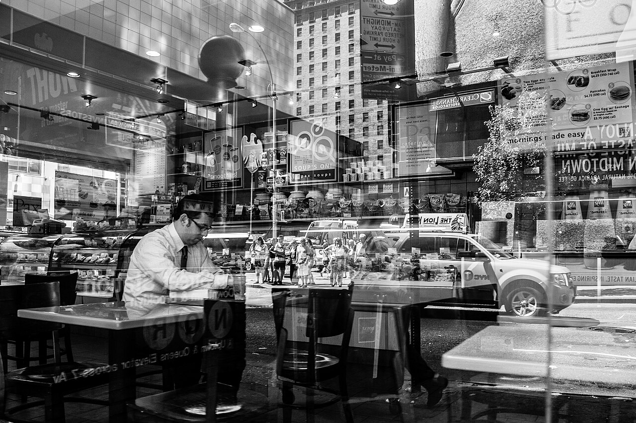 A man dining alone beside a Manhattan restaurant window, seen through layered reflections of the room and street.