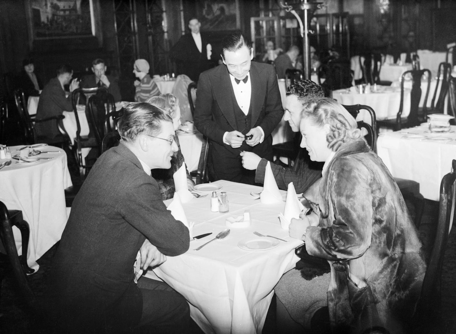 A waiter takes an order from four diners at linen-covered tables in a London West End restaurant in spring 1941.