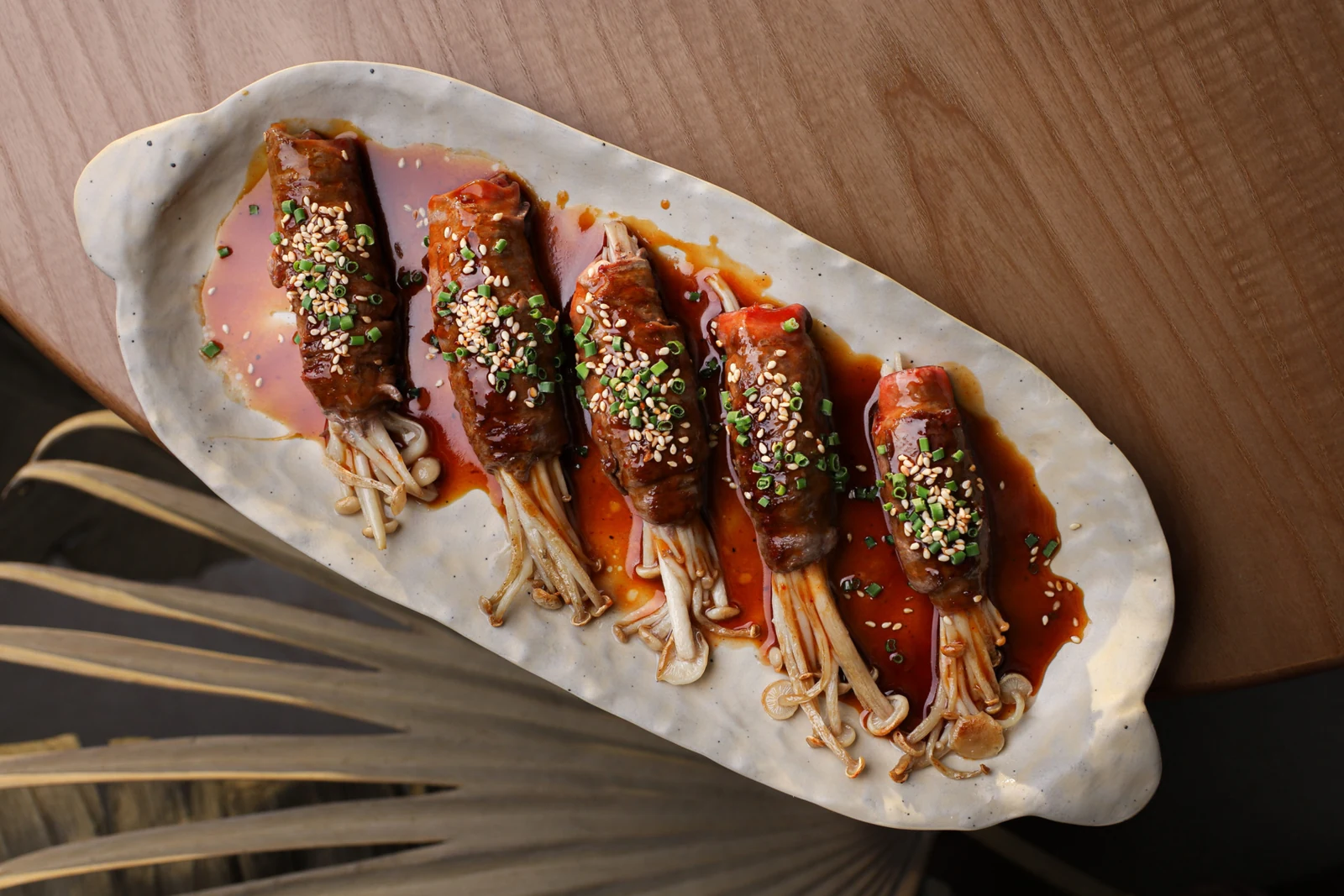 Five bundles of enoki mushrooms wrapped in glazed veal, arranged across a long ceramic plate with sesame seeds and chopped scallions.