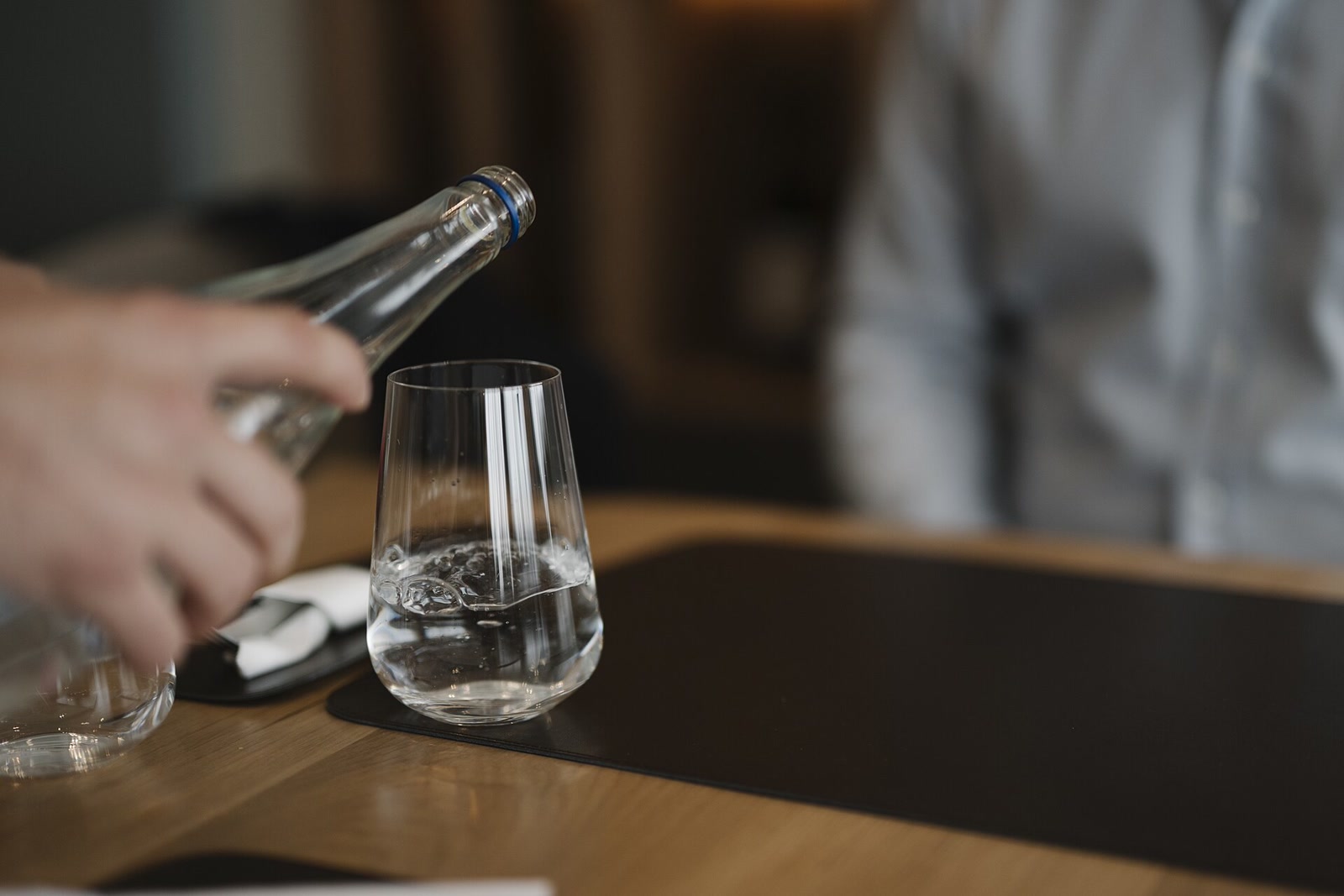 A waiter's hand pours water from a clear bottle into a drinking glass at a warmly lit restaurant table.