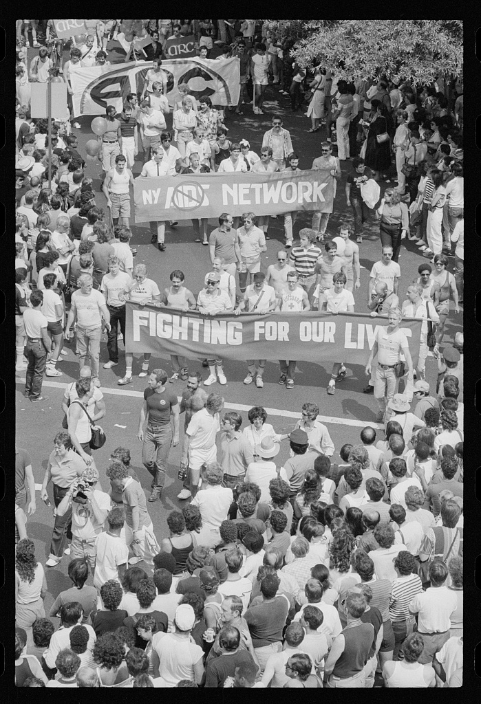An overhead archival photograph of a crowded 1983 New York march, with participants carrying NY AIDS Network and Fighting for Our Lives banners.