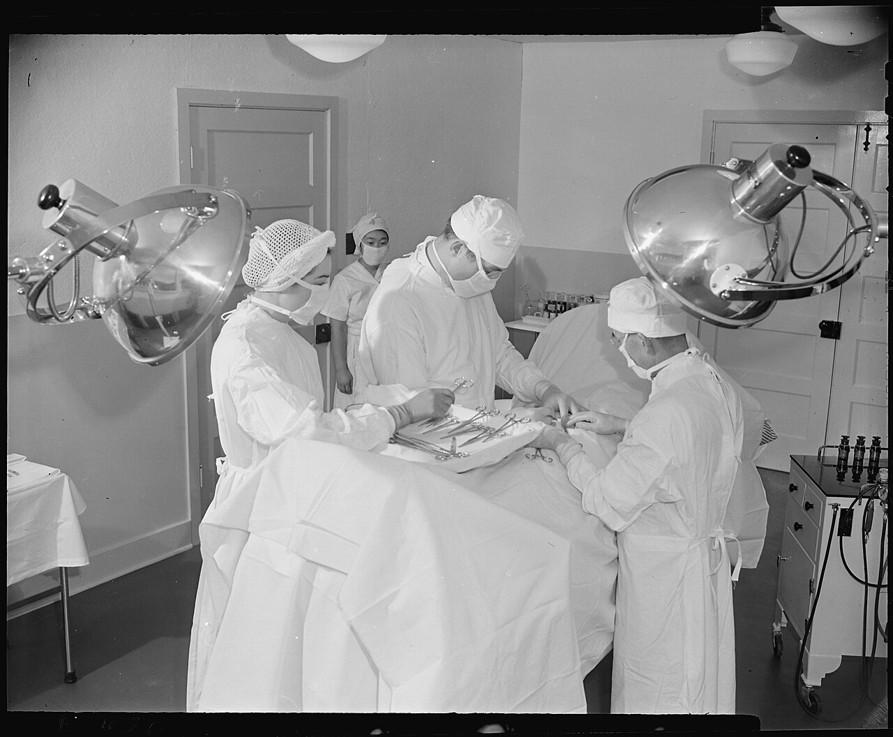 Four masked clinicians perform an emergency appendectomy beneath two large surgical lamps in the Amache hospital operating room in 1942.