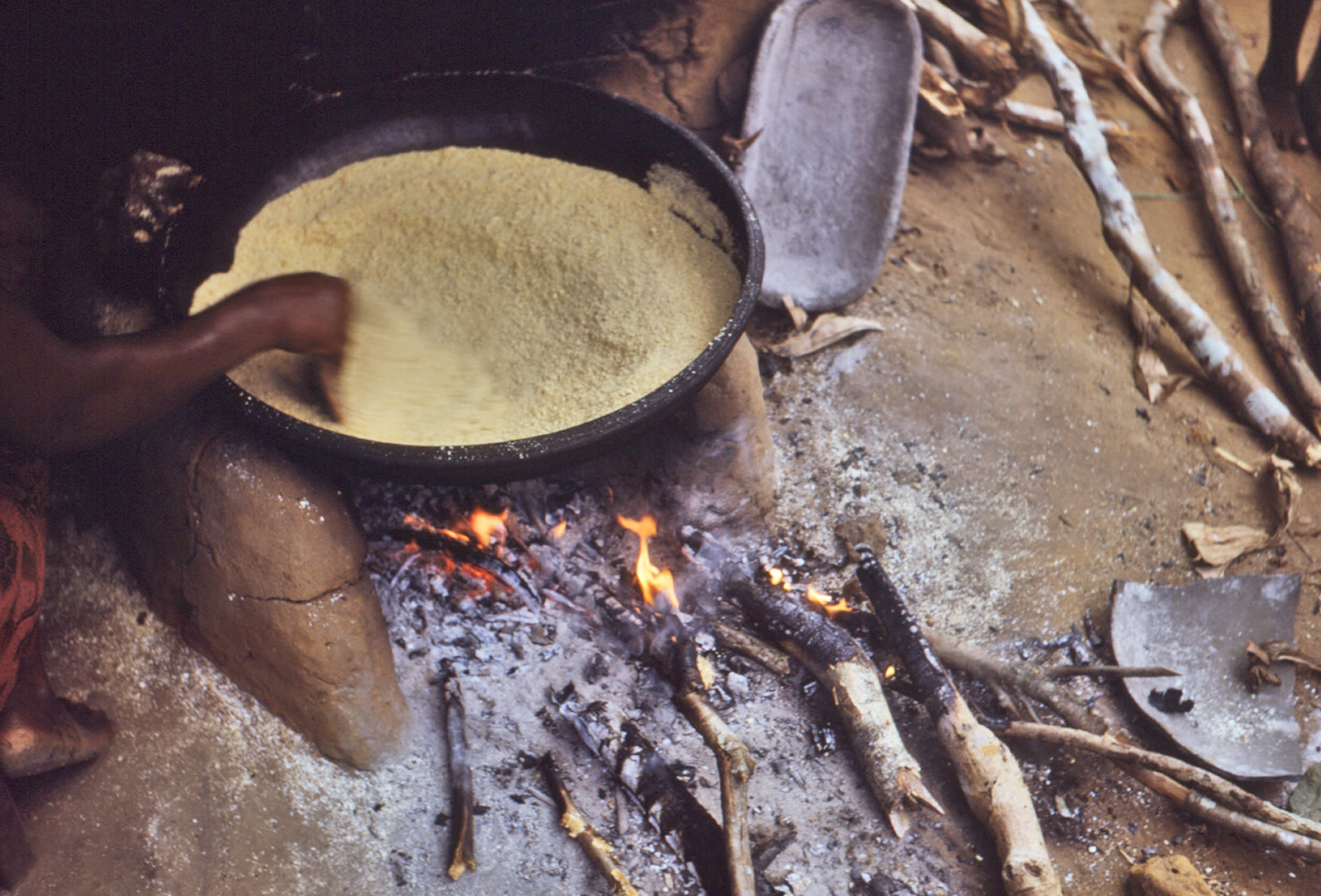 A hand stirs pale corn-soya food in a wide black pan over a wood fire at a Nigerian-Biafran refugee camp in 1968.