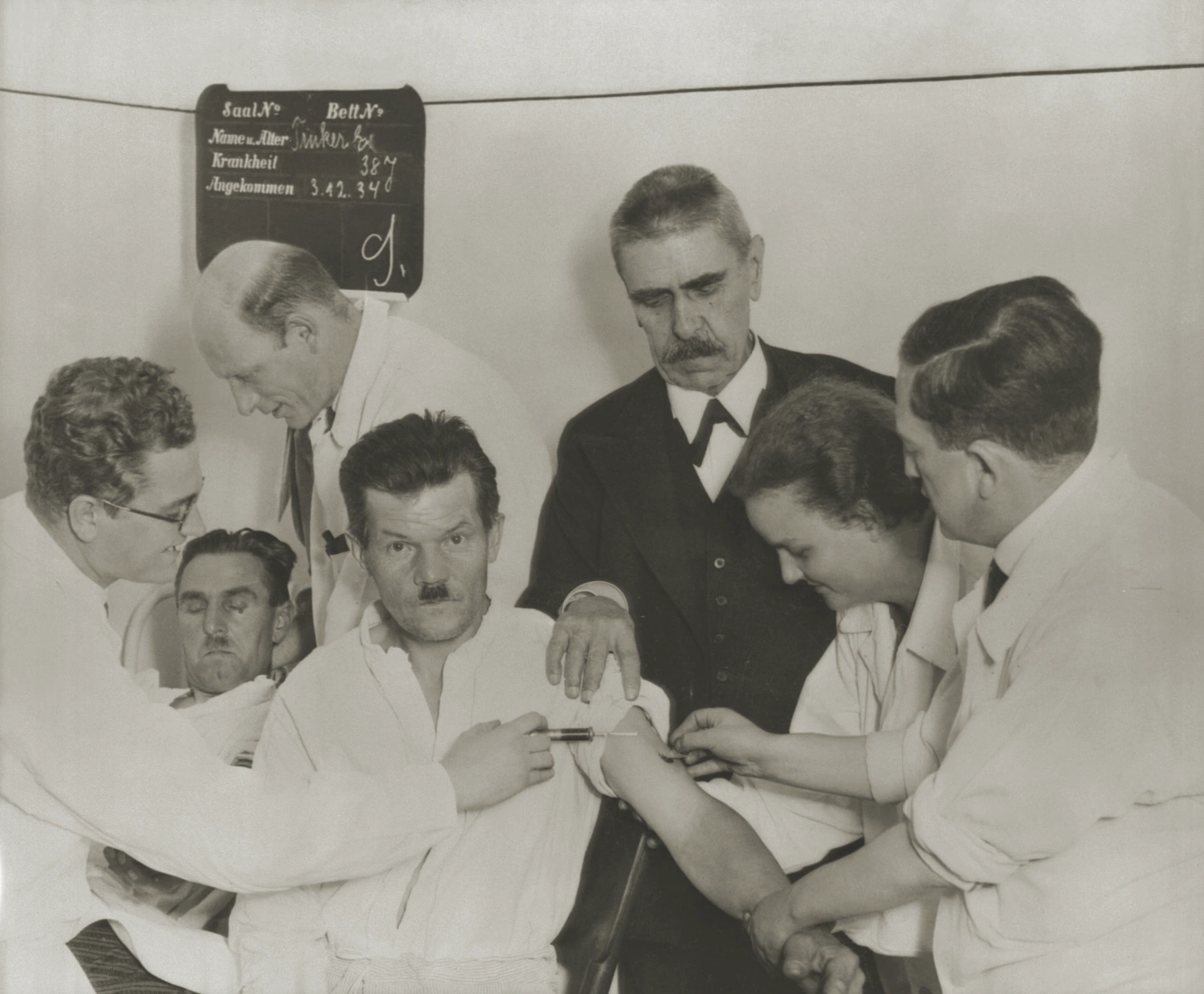 Black-and-white 1934 clinical photograph of staff transferring blood from a malaria-infected patient to a seated patient with neurosyphilis while Julius Wagner-Jauregg watches.