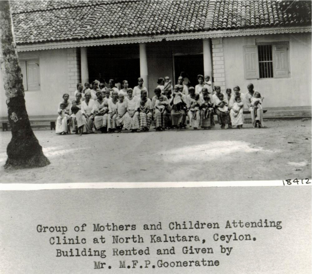 Black-and-white archival photograph of mothers holding children outside a tiled-roof clinic in North Kalutara, Ceylon.