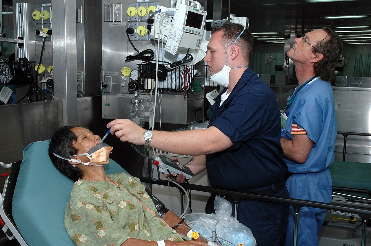 A Navy hospital corpsman holds a clinical thermometer at a seated patient's mouth while a nurse monitors other vital signs aboard USNS Mercy.
