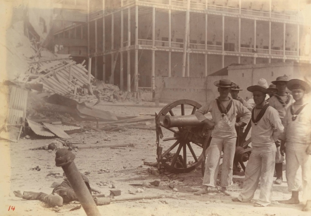 Archival sepia photograph of British marines posing around a captured Zanzibari cannon amid palace wreckage, with two motionless bodies visible at lower left.