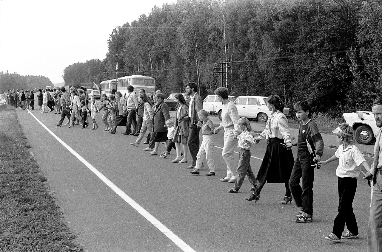 Black-and-white archival photograph of Baltic Way participants standing hand in hand along the Vilnius-Ukmergė motorway, with buses and cars parked beside the road.