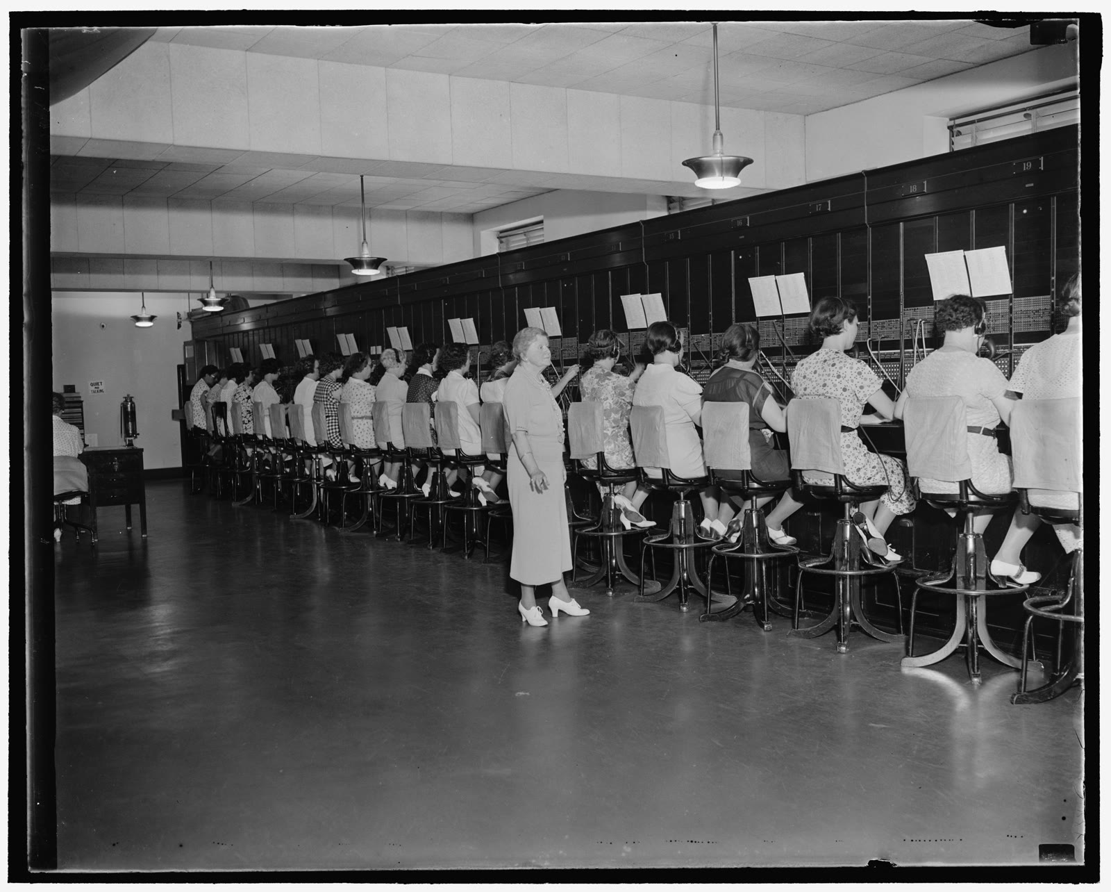 1937年的黑白档案照片：哈里奥特·戴利站在一排排女电话话务员旁，她们在美国国会大厦的人工交换台工作。