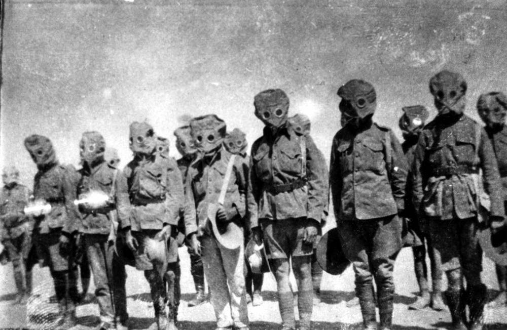 Grainy black-and-white archival photograph of soldiers standing outdoors in a line while wearing hood-like First World War gas masks.