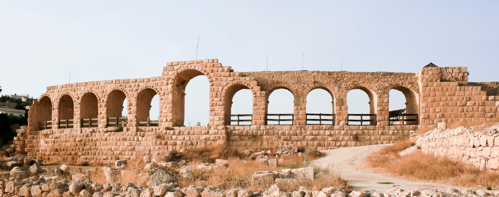 Panoramic daylight photograph of the arched stone end of the ancient Roman hippodrome at Jerash, with rubble and dry grass in the foreground.