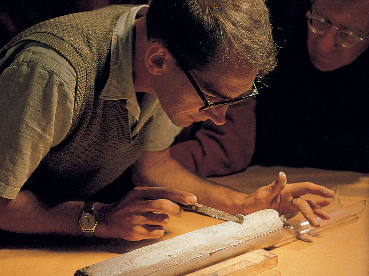 Color archival photograph of oceanographer William Riedel leaning over a cylindrical sediment core recovered during Project Mohole in 1961.