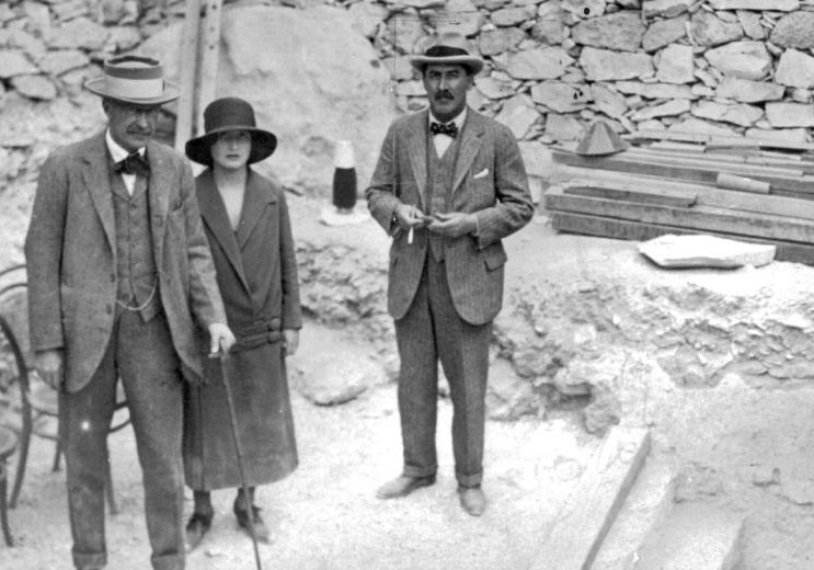 Black-and-white archival photograph of Howard Carter, Lord Carnarvon, and Lady Evelyn Herbert standing at the stone steps leading to Tutankhamun’s newly discovered tomb in November 1922.
