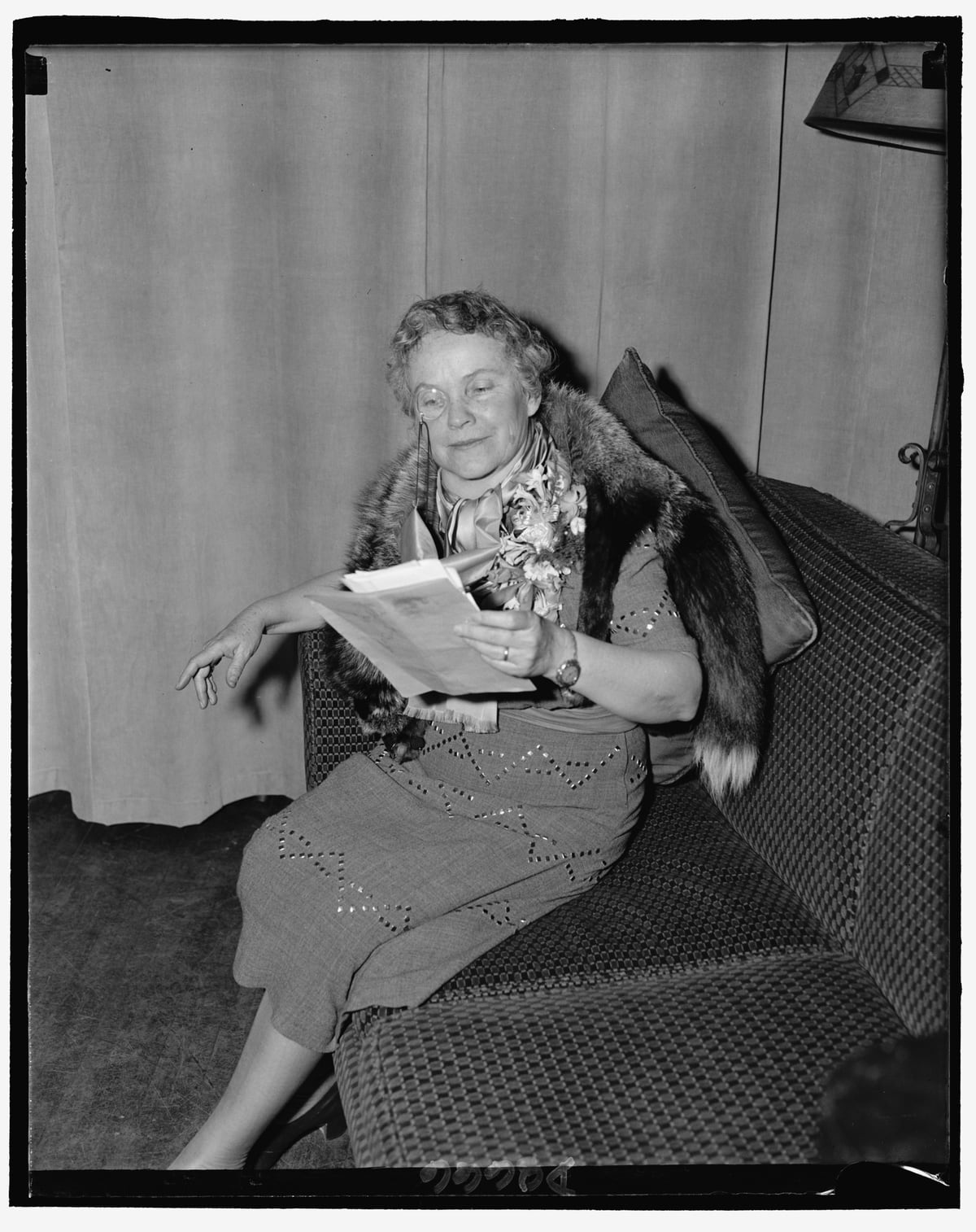 Black-and-white archival portrait of Dorothy Canfield Fisher seated on a patterned sofa, holding papers with her gaze lowered to one side.
