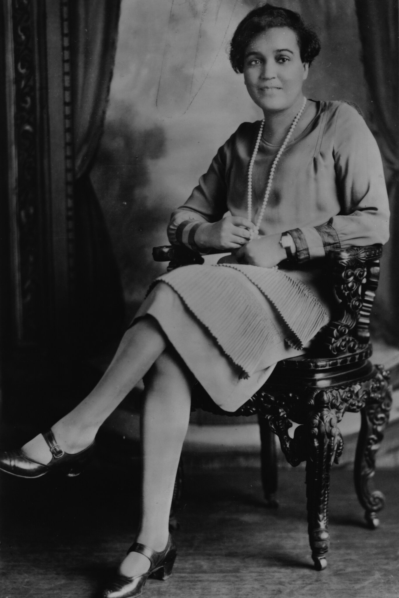 Black-and-white studio portrait of Jessie Redmon Fauset seated in a carved chair, wearing a long strand of pearls and looking directly at the camera.