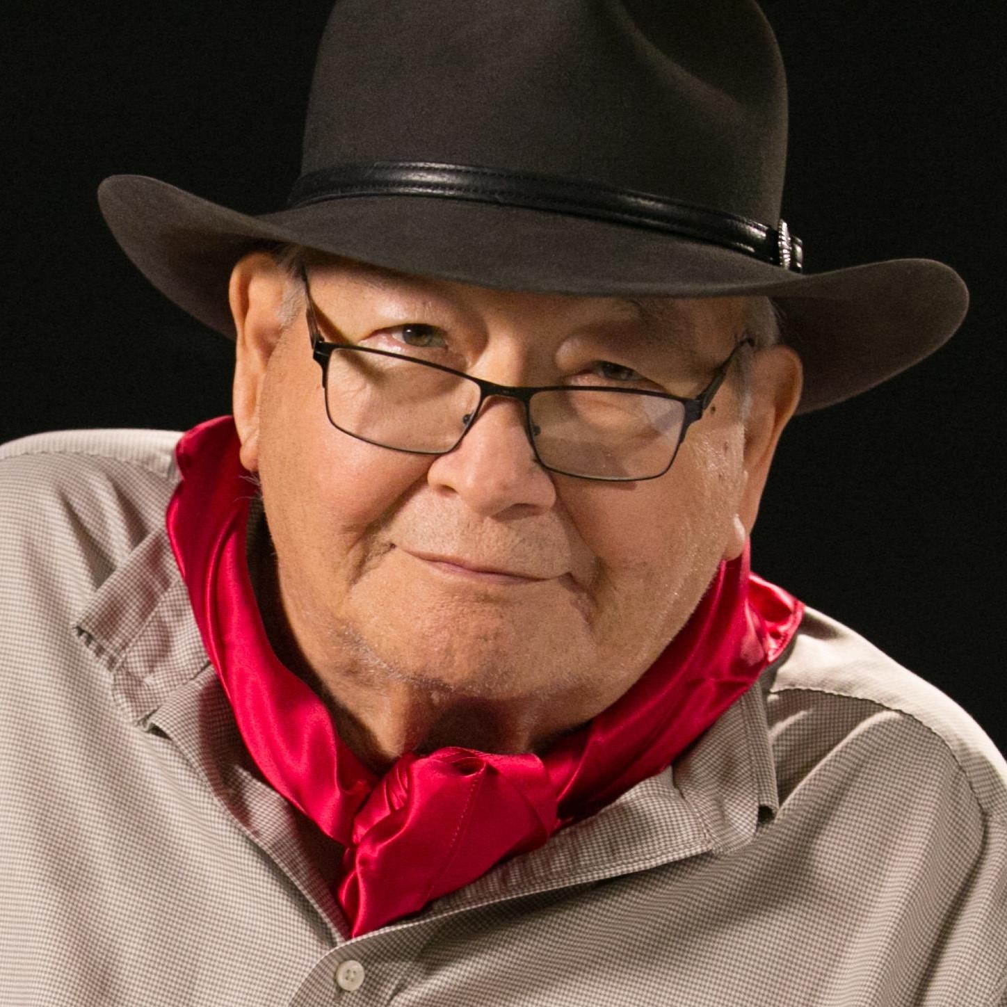 N. Scott Momaday in a close photographic portrait wearing glasses, a broad dark hat, checked shirt, and bright red neckerchief against a black background.