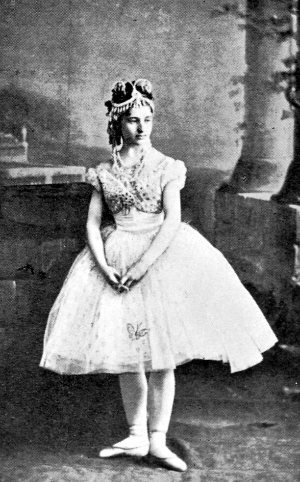 Full-length black-and-white archival studio photograph of Giuseppina Bozzacchi standing with her hands lightly clasped, costumed as Swanilda in the original 1870 production of Coppélia.