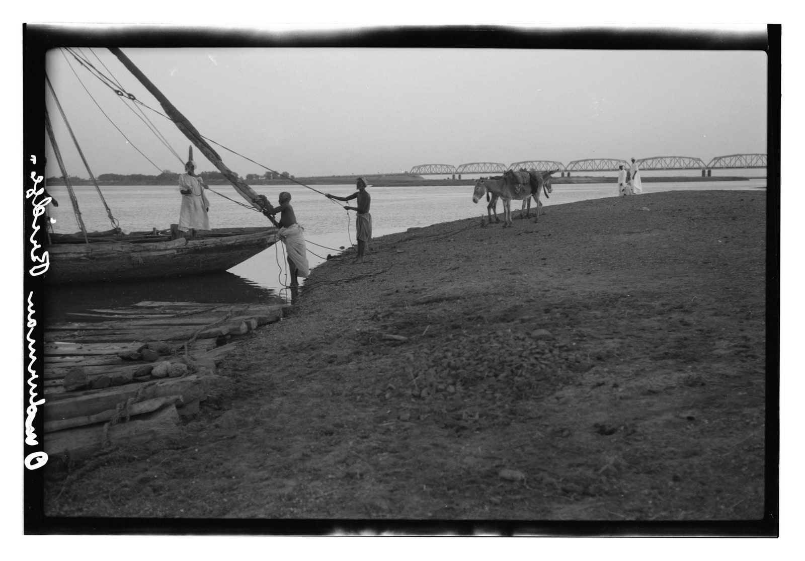 Black-and-white archival photograph of men working beside a wooden boat on the Nile, with pack animals on the bank and Omdurman Bridge in the distance.