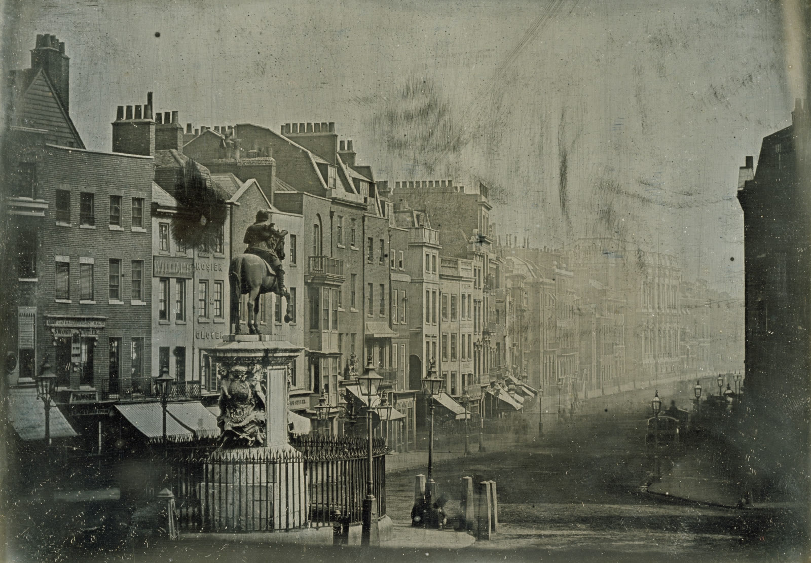 Early monochrome photograph looking down Whitehall from Trafalgar Square, with an equestrian statue, rows of buildings, and traffic blurred into ghostlike traces.