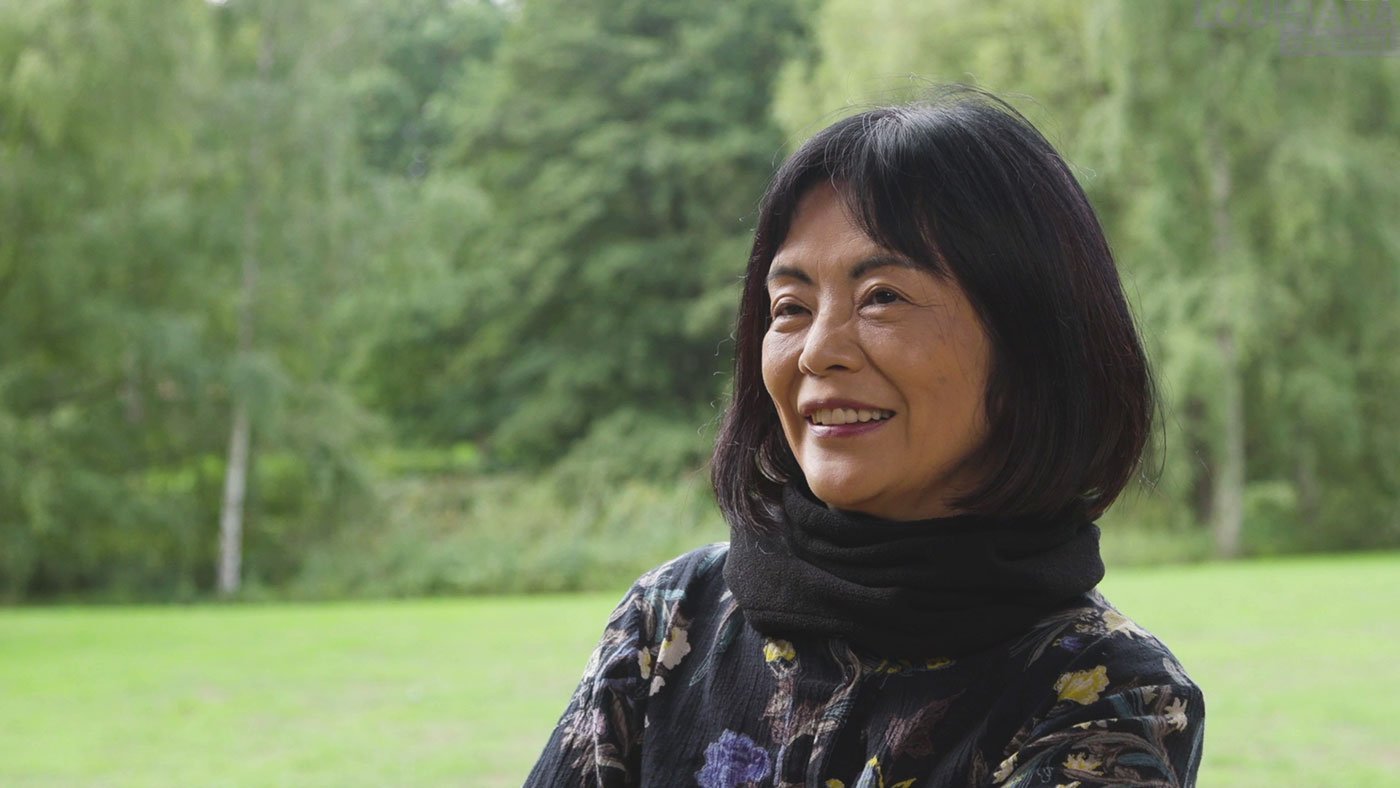 Yoko Tawada seated outdoors during her 2018 Louisiana Literature interview, with green trees and lawn behind her.