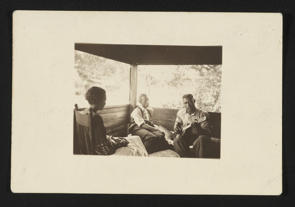 Black-and-white archival photograph of Zora Neale Hurston seated with Rochelle French and guitarist Gabriel Brown on a screened porch.