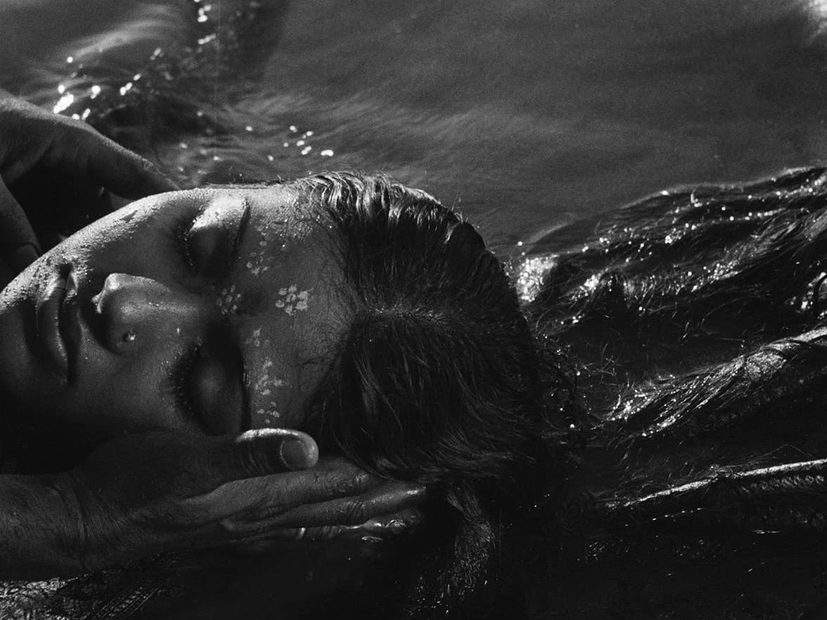 Black-and-white film still from A River Called Titas showing a woman floating at the river's surface while hands steady her head.