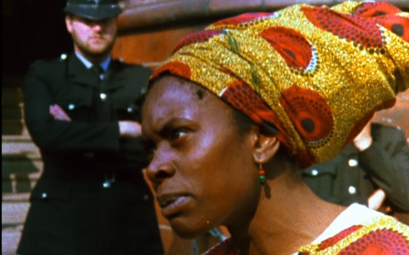Colour film still from Handsworth Songs showing a Black woman in a patterned headwrap in the foreground with uniformed police officers behind her.