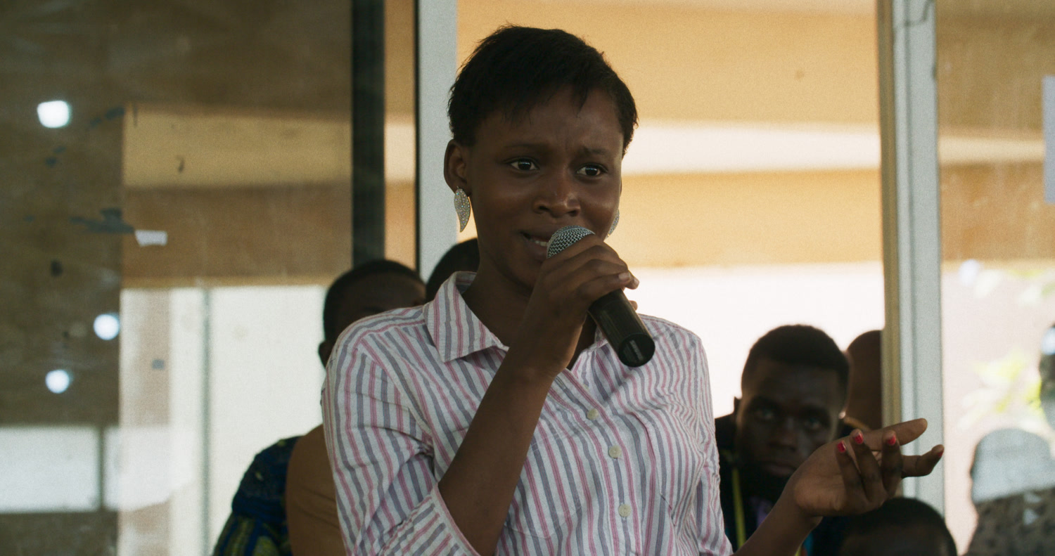 A young woman in a striped shirt speaks into a handheld microphone during the University of Abomey-Calavi debate in Dahomey.