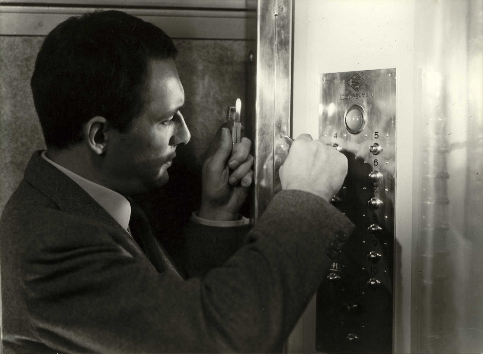 Black-and-white official still from Elevator to the Gallows showing Maurice Ronet as Julien Tavernier holding a lighter beside the elevator controls.