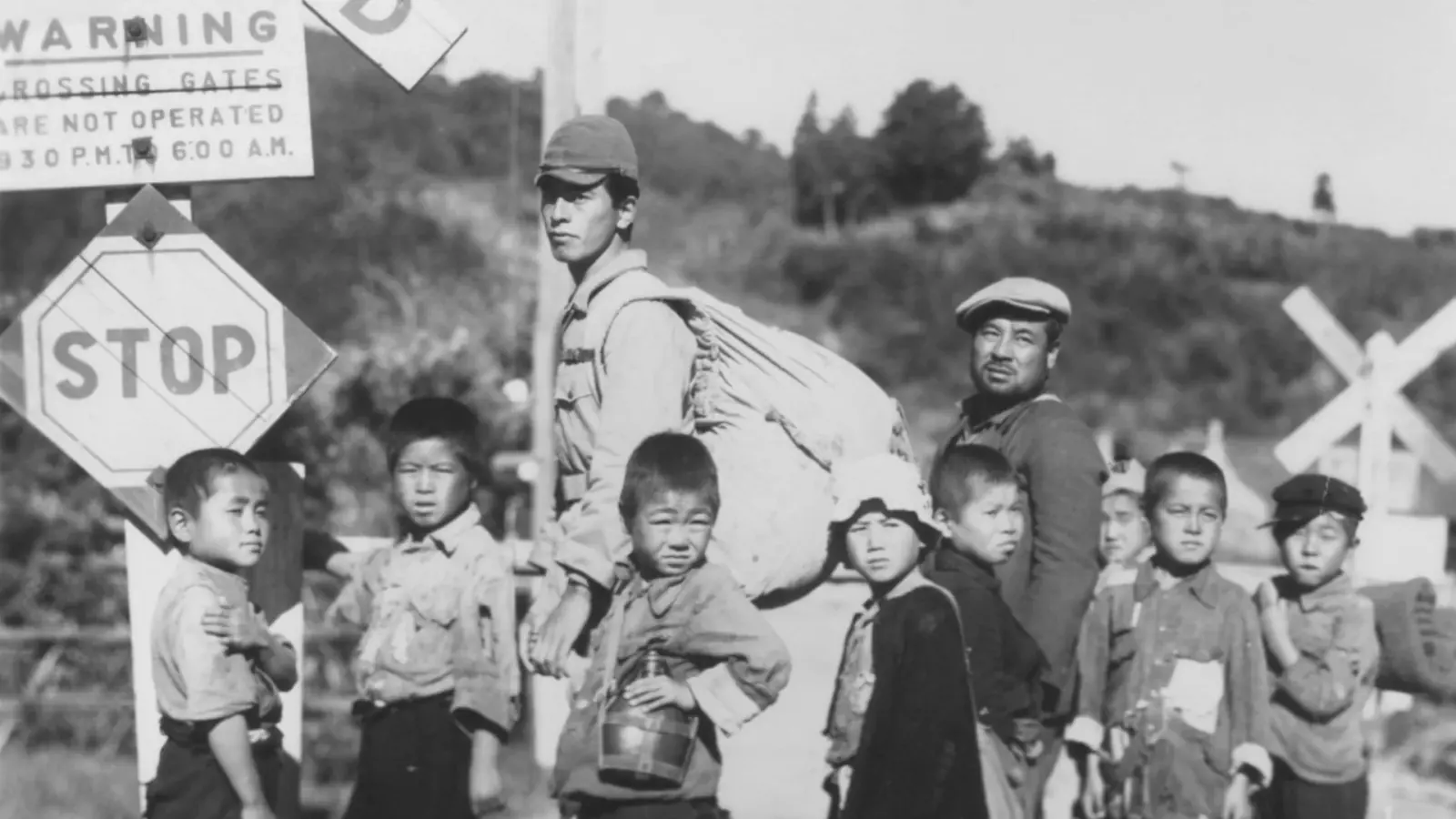 Black-and-white film still of two men and eight boys at a rural railway crossing, with one man carrying a large bundle.