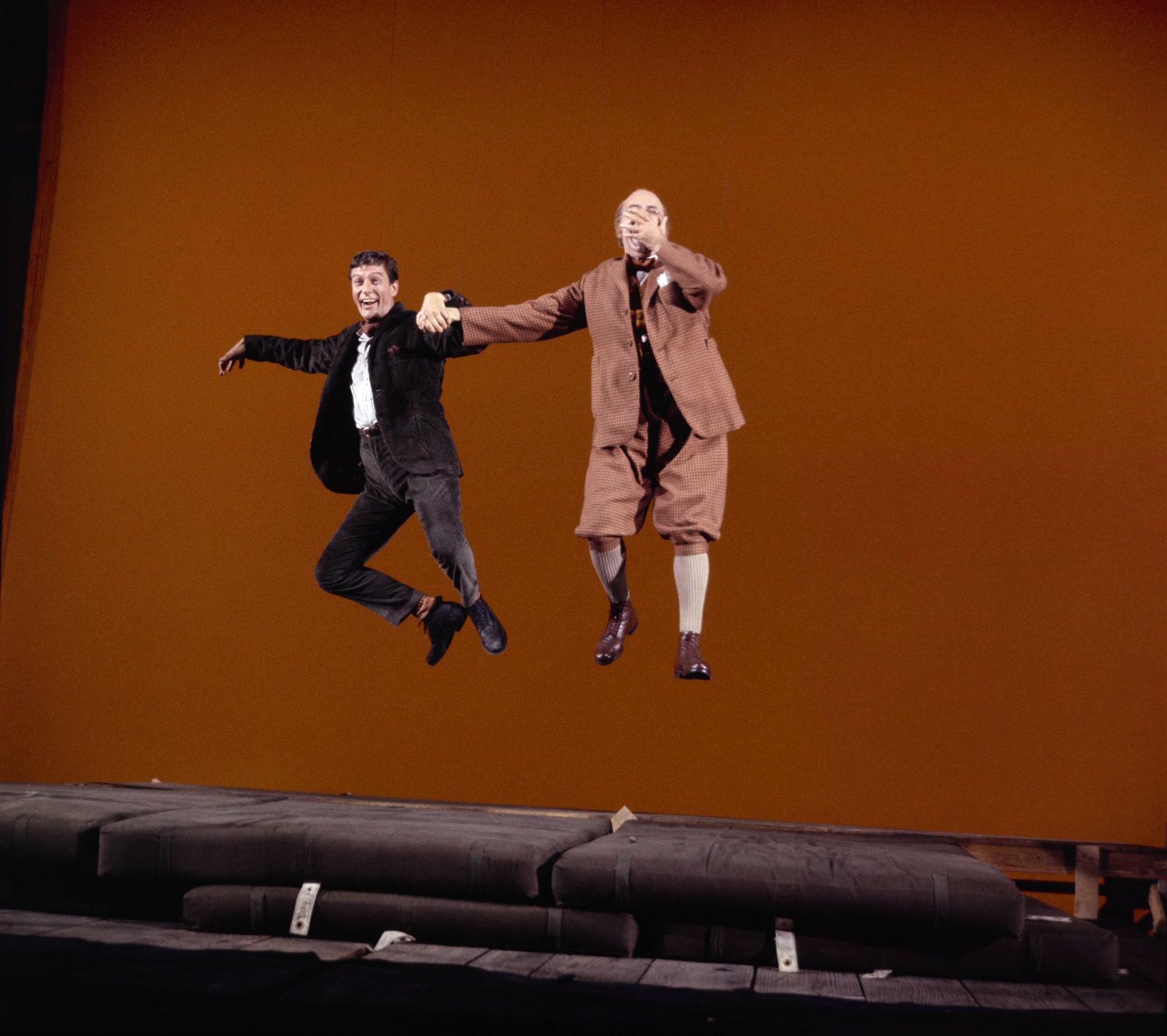 Archival production photograph of Dick Van Dyke and Ed Wynn suspended above crash mats in front of a plain amber backing on the set of Mary Poppins.
