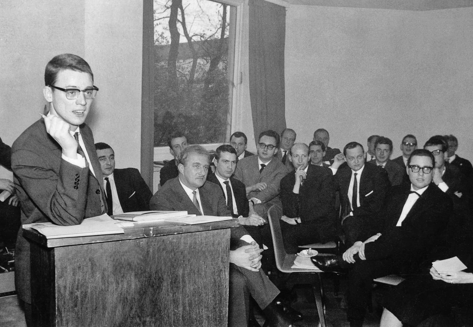 Alexander Kluge speaking at a lectern before a room of Oberhausen Manifesto signatories at the declaration's 1962 press conference.