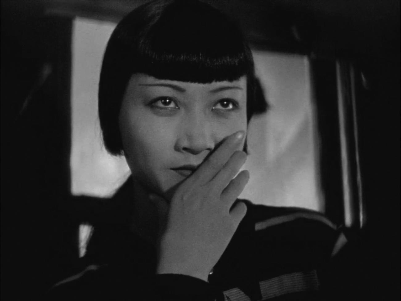 Archival still from the 1929 film Piccadilly showing Anna May Wong as Shosho, her face turned toward the camera with one hand raised beside it.
