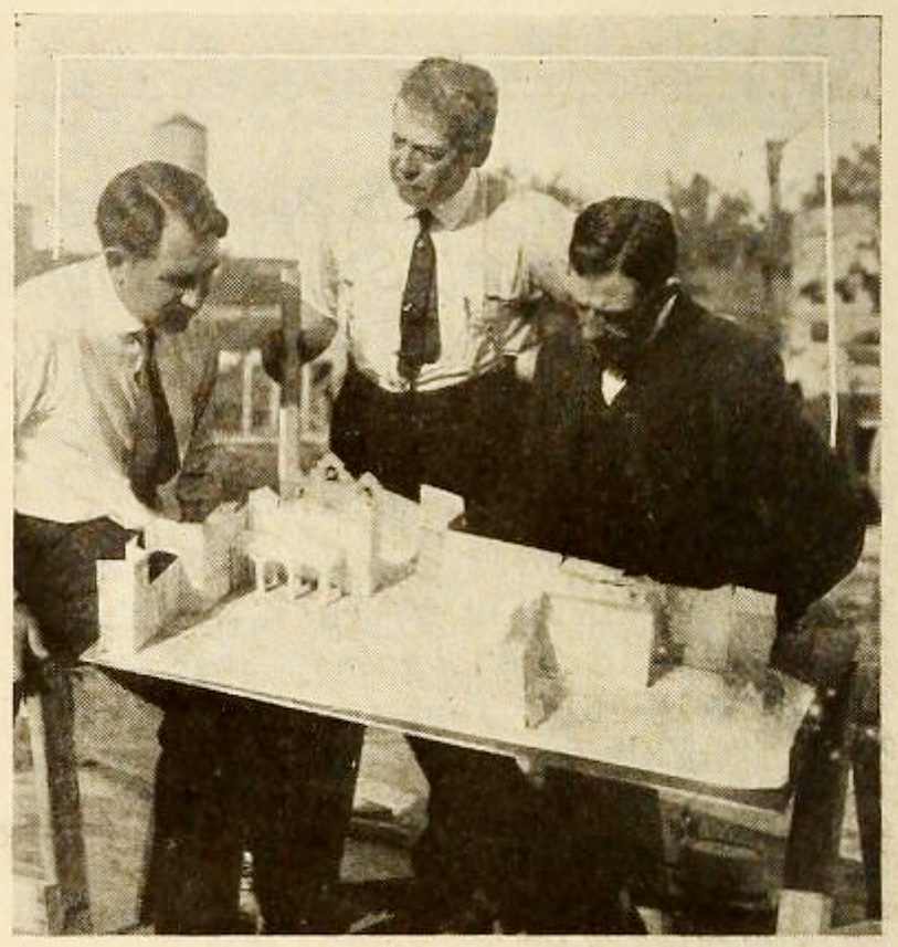 Three film workers lean over a tabletop architectural model of a village in a 1915 archival photograph.