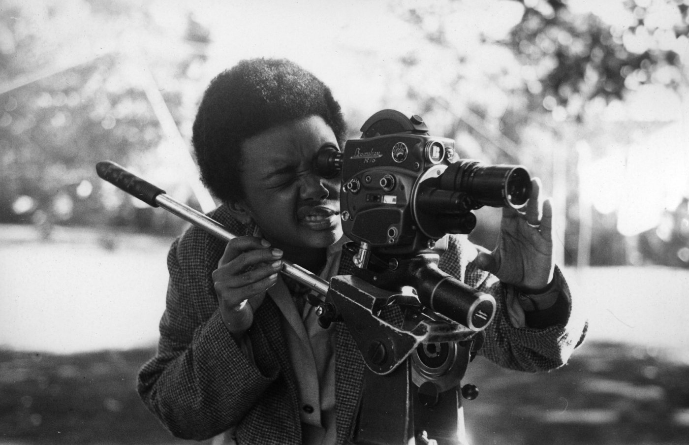 Black-and-white archival photograph of Sara Gómez leaning into the viewfinder of a motion-picture camera outdoors.