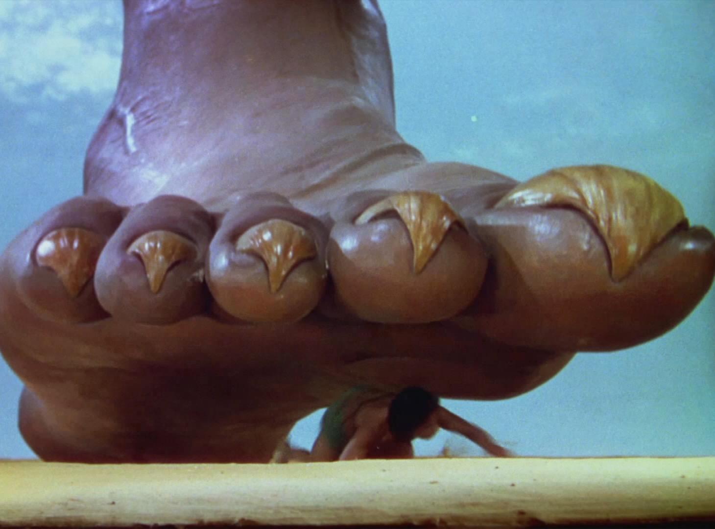 Color film frame showing Sabu as Abu crouching beneath the enormous bare foot of Rex Ingram's Djinni against a blue sky.