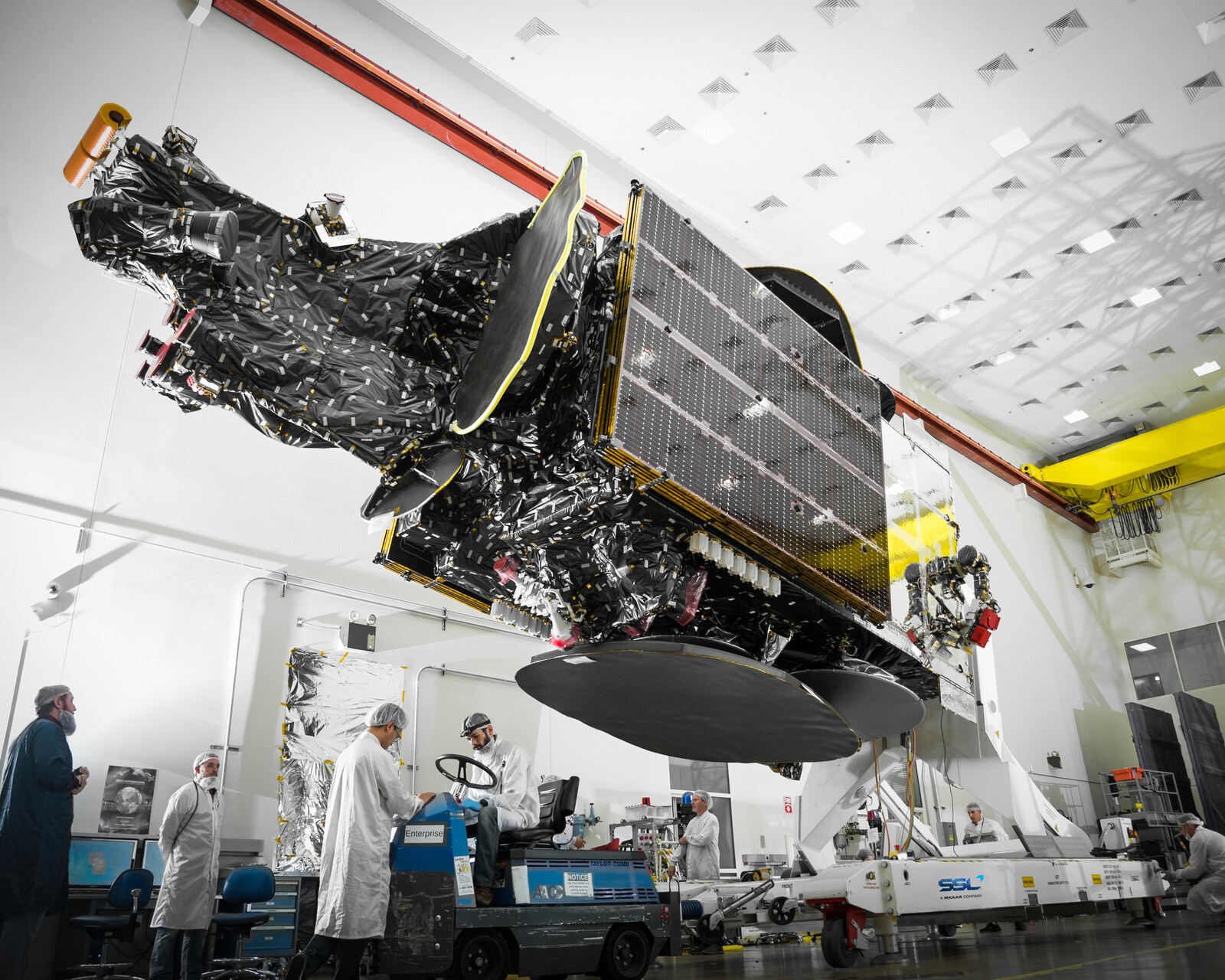 Engineers in cleanroom clothing work beneath the large Telstar 18 VANTAGE communications satellite in a white high-bay facility.