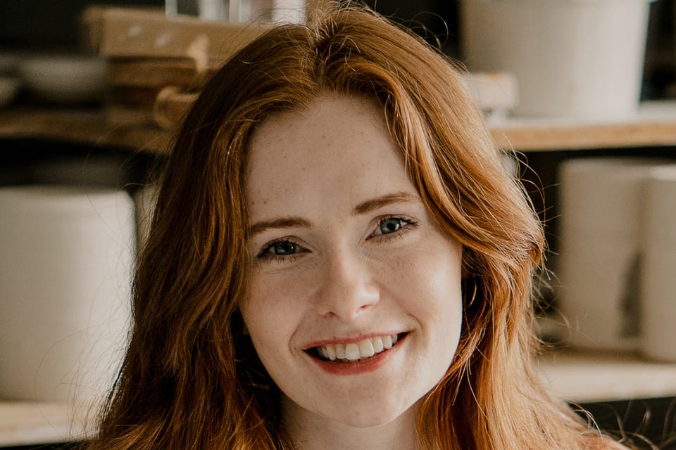 Potter Natasha Patterson smiles in her ceramics studio, with shelves and ceramic vessels behind her.
