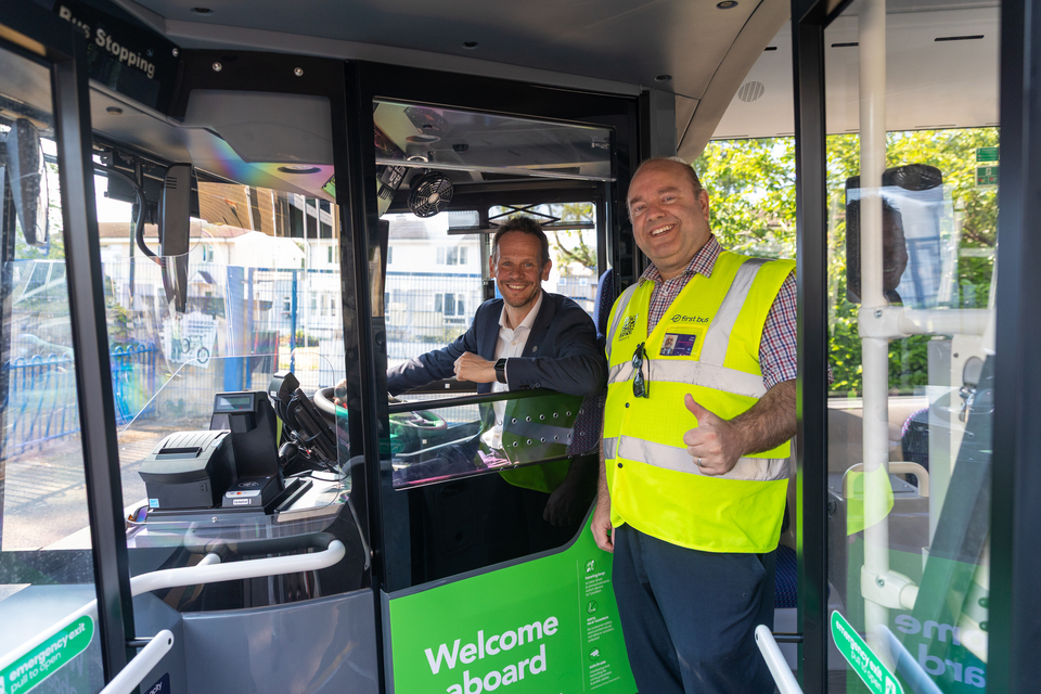 A suited transport official and a First Bus driver smile from the entrance of a green bus.