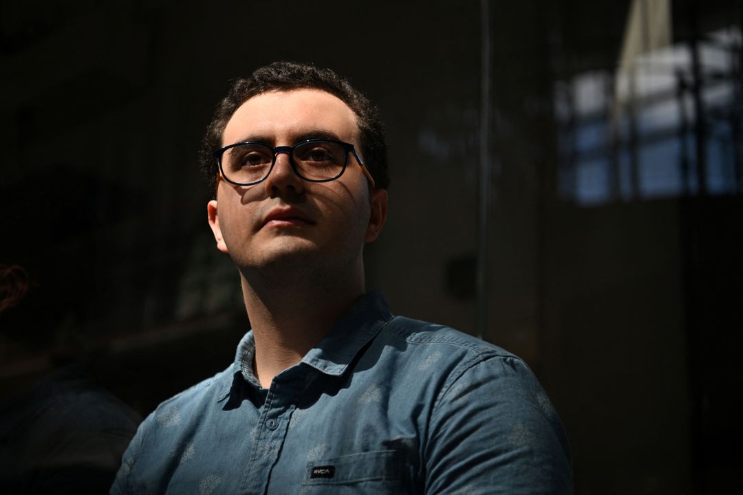 Sinan Can Demir, wearing glasses and a blue shirt, poses beside dark reflective glass in Austin.