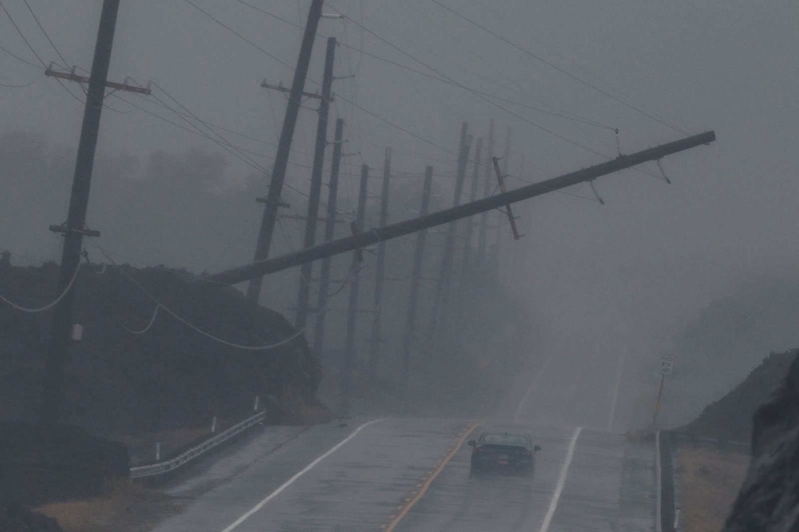 A car drives through heavy rain near a downed utility pole dangling above Māmalahoa Highway near Ocean View on Hawaiʻi Island.